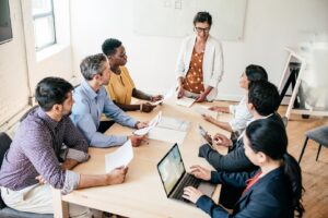 A woman stands at the head of a table while colleagues sit around the table smiling