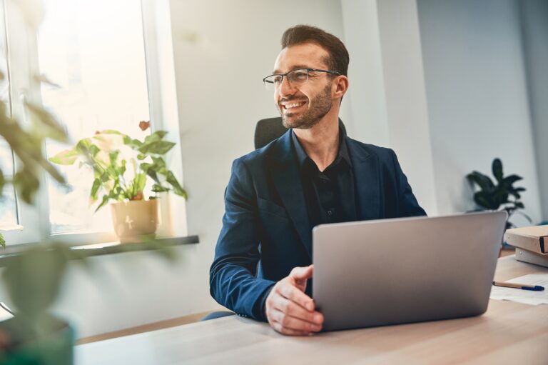 a man with glasses holds a laptop on his desk and smiles