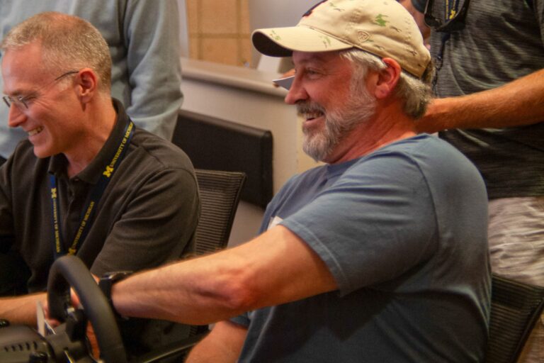 A man in a ball cap and t-shirt smiles while turning a steering wheel in a room