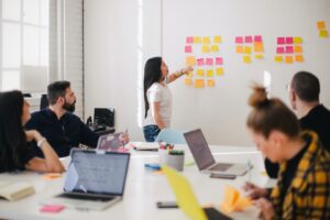 A woman points at sticky notes on a white board while her teammates sist around a table and listen