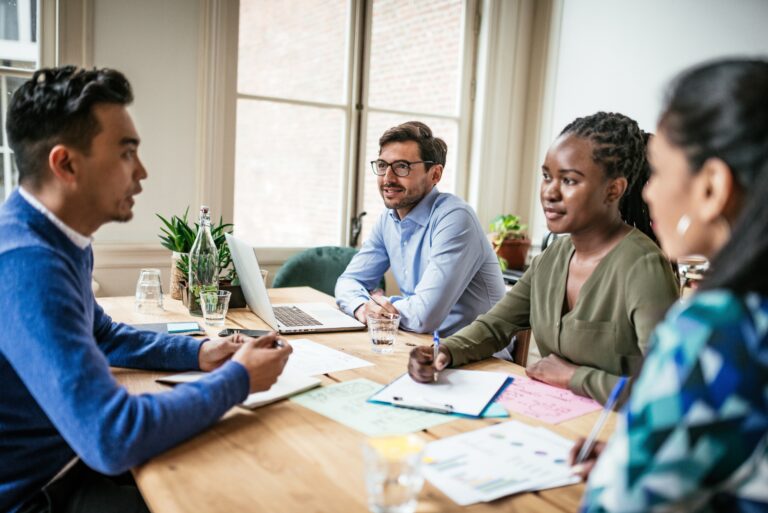 Two men and two women sit around a table with papers, pens, and a laptop