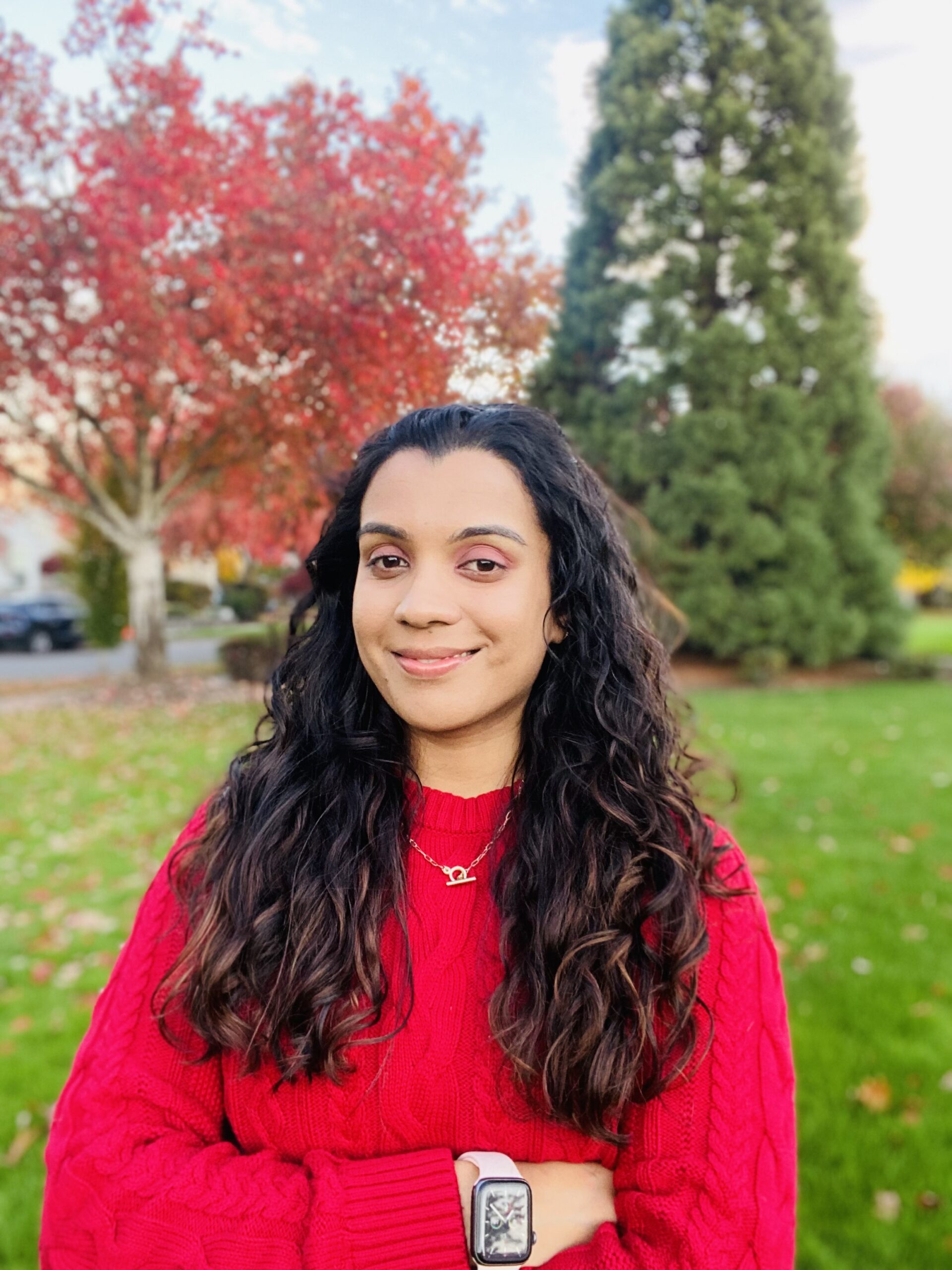 A woman with long, brown curly hair wears a red sweater and smiles in front of trees changing colors