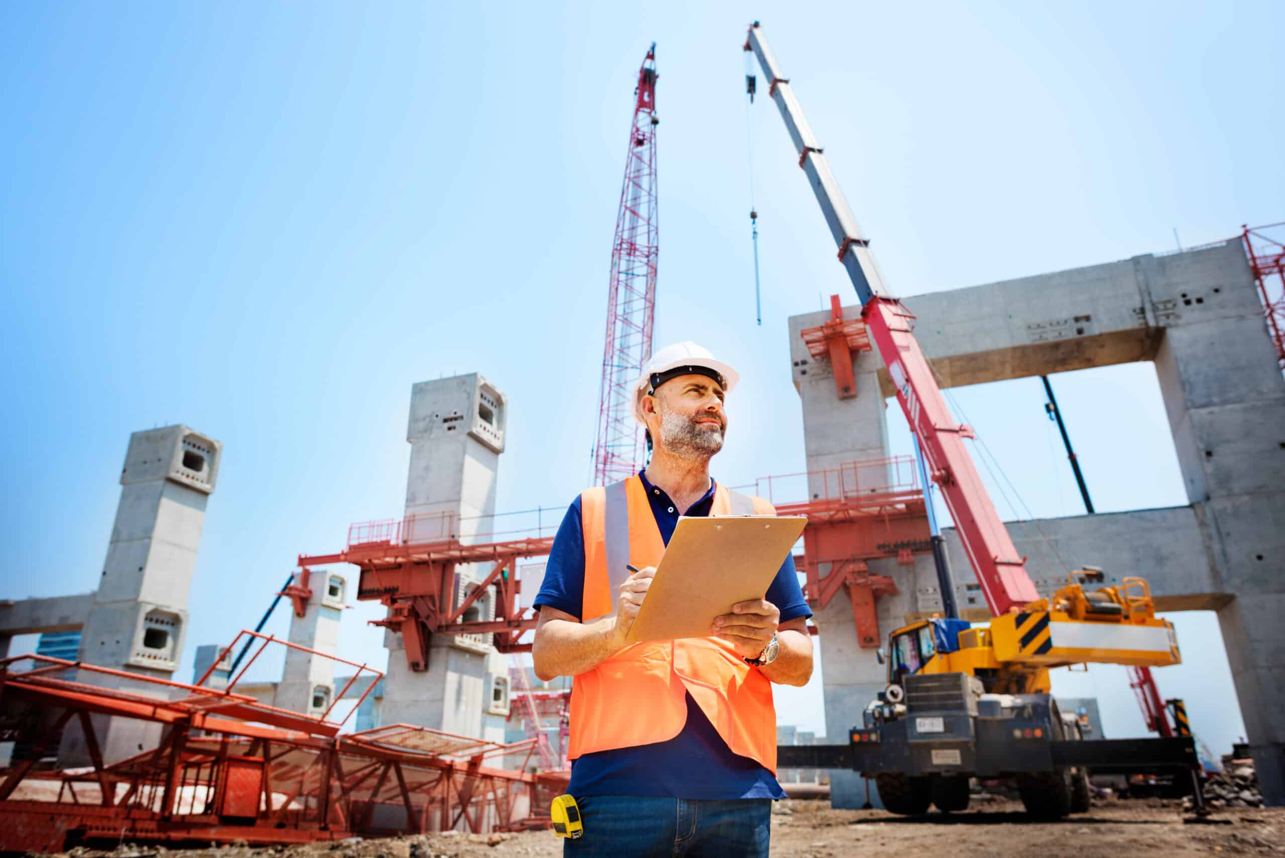 A man in an orange vest holding a clipboard while standing in front of a construction site.
